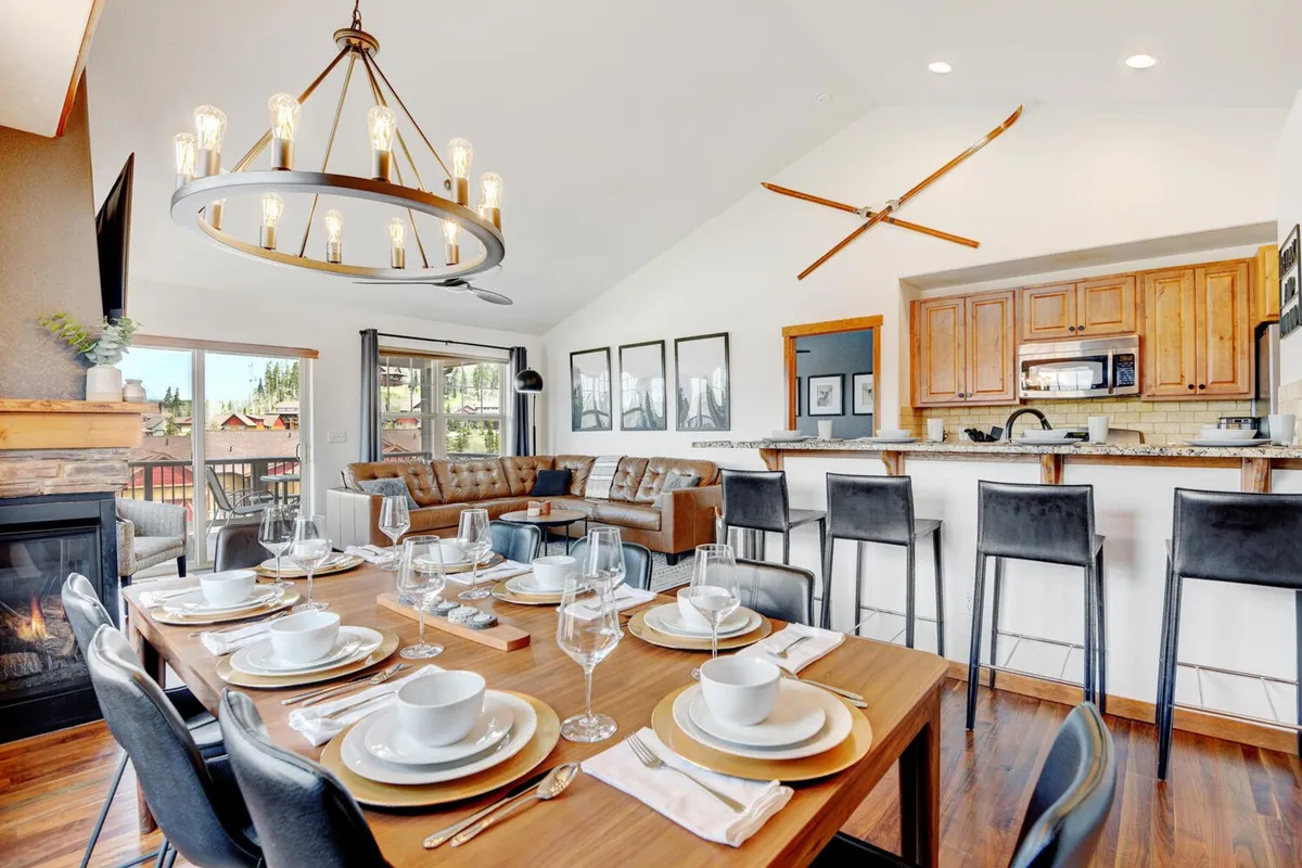 A warm and welcoming interior view showcasing a dining table set for a meal, a glowing fireplace, a comfortable couch, and a visible kitchen area behind. The chandelier adds an elegant touch, and large windows bring in mountain light. The space feels ready for family dinners, holidays, or cozy nights in.