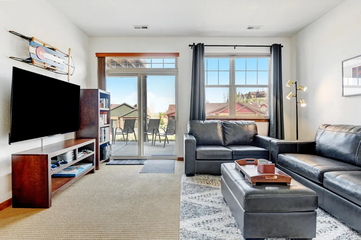 A cozy family room featuring a black leather sofa, a wooden TV unit with a large television, expansive windows offering mountain views, and a bookshelf filled with books. The overall layout invites relaxation, movie nights, and shared family activities.
