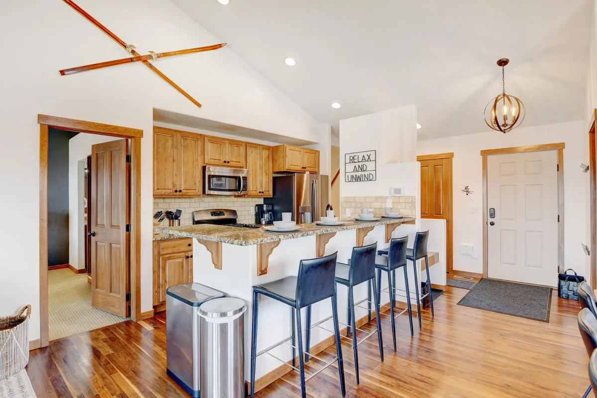 A modern kitchen with a central island, stylish bar stools, and rich wooden cabinetry. A sign reading 'RELAX AND UNWIND' adds a personal, welcoming touch. The kitchen is equipped for both casual brunches and elegant dinners, inviting guests to cook, gather, and linger.