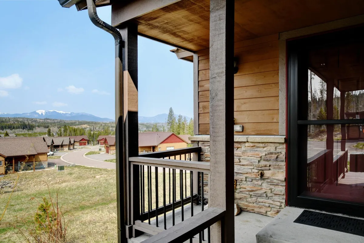 A wide porch with wooden railings and stone accents overlooks a breathtaking mountain view. The scene captures the inviting exterior architecture and the tranquil natural surroundings, suggesting a peaceful place to relax or work with inspiration from the outdoors.