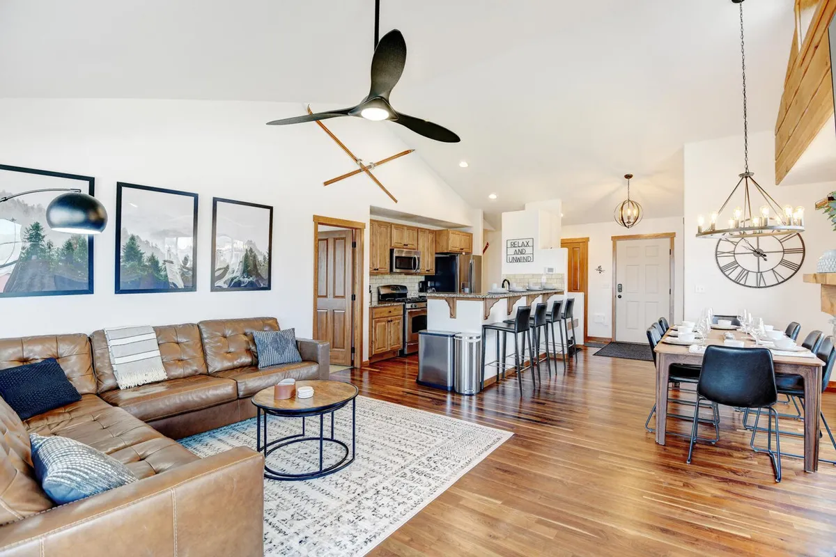 A spacious, open-concept living area featuring a plush leather sofa, a wooden dining table with seating for eight, and a modern kitchen island. The decor includes inviting wall art and large windows that flood the space with natural light. The layout fosters seamless movement between the living room, dining area, and kitchen, ideal for gatherings and entertaining.