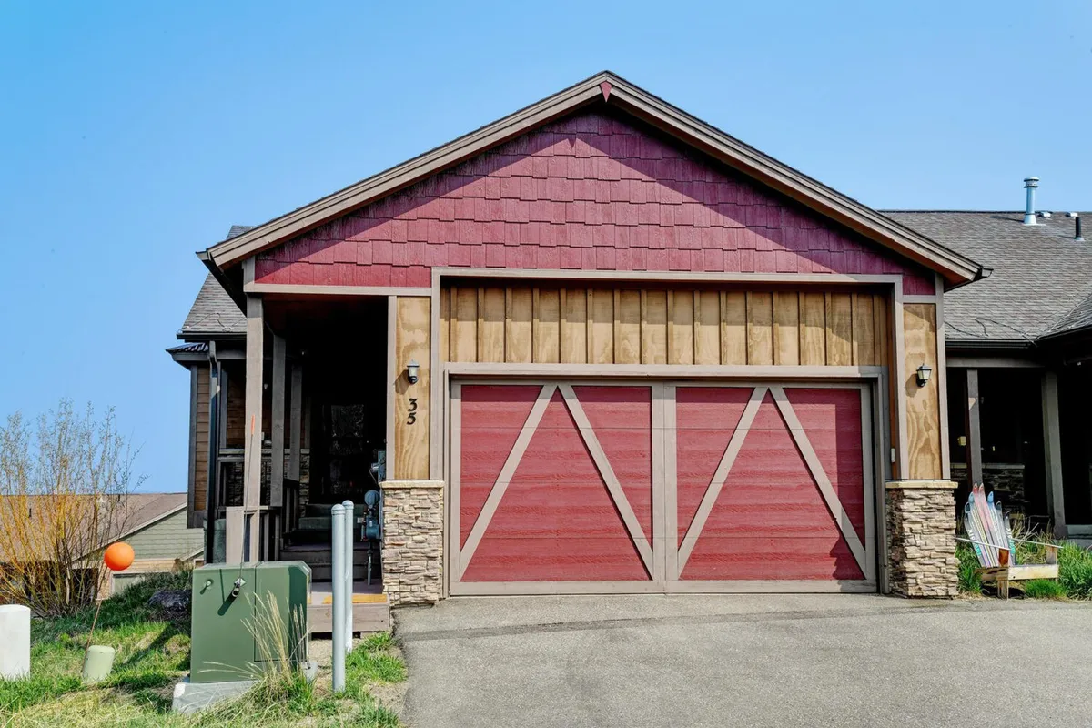 The image shows the front view of the home with its distinctive red garage door, stone accents, and warm wooden siding. The scene is welcoming, with plenty of space in front of the house for outdoor play or gatherings. The mood is friendly and inviting, suggesting a place where families can enjoy time together outdoors.