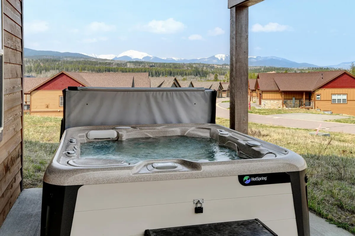 A steaming outdoor hot tub set against a dramatic mountain backdrop, framed by a wooden structure. The scene exudes relaxation and offers a glimpse of the natural adventure available right outside the door.