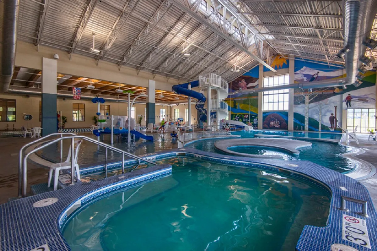 This vibrant interior scene at a recreation center features lively children and families enjoying water slides and swimming pools, set against colorful murals and ample poolside seating. The joyful, energetic atmosphere highlights communal activities and recreational fun.