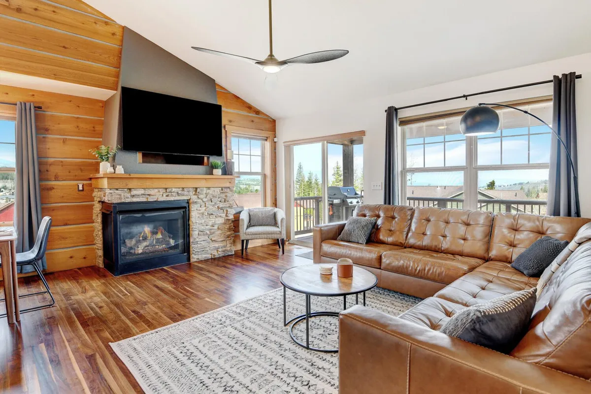 This inviting interior scene showcases a spacious living area with a cozy stone fireplace, plush leather sofa, and expansive windows framing mountain views. The coffee table at the center offers a perfect gathering spot, while the natural light floods the room, creating a warm, welcoming atmosphere ideal for relaxation or entertaining after a day of adventure.