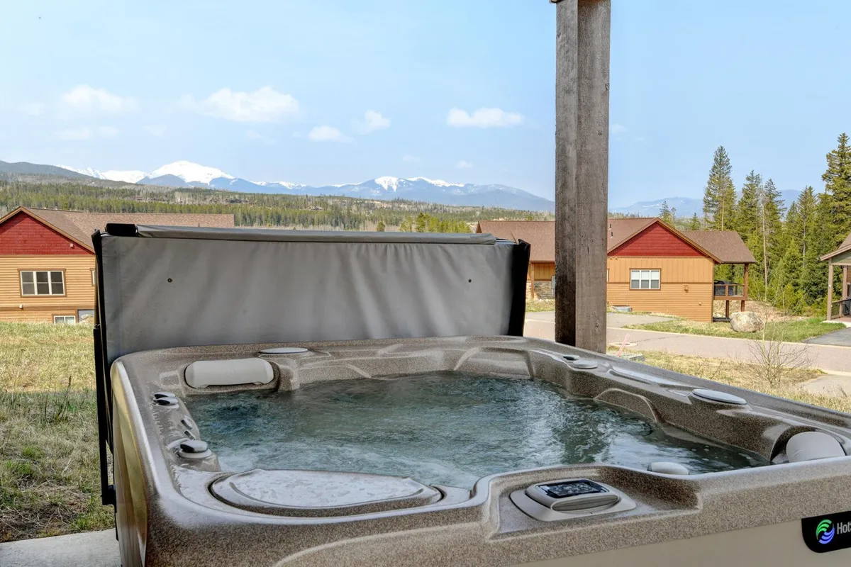 An exterior scene showing a luxurious hot tub set against a breathtaking mountain backdrop. The surrounding landscape features natural foliage and the sky glows with soft sunset hues, inviting relaxation and social gatherings.